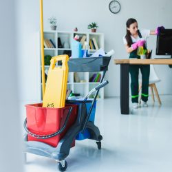 young female janitor cleaning office with various cleaning equipment